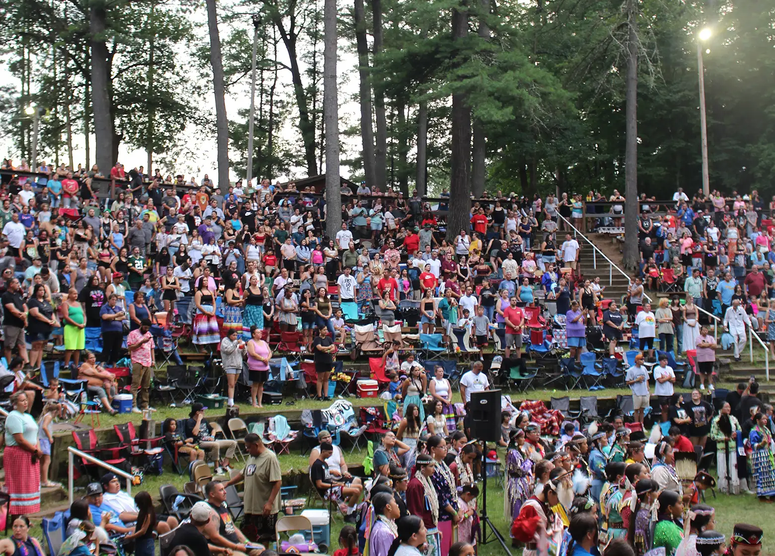 Large crowd gathered outdoors in a forested amphitheater, many in traditional Native regalia, attending a powwow.