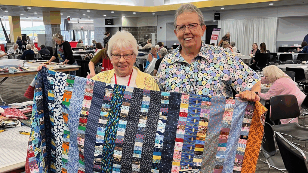 Two women smile holding up a quilt in an expo center meal space, people at tables and chairs behind them.