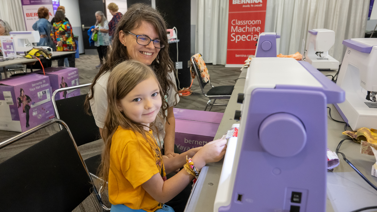 A woman and a young girl work together at a sewing machine in an expo center, a sign behind them reads, "BERNINA Classroom Machine Special."