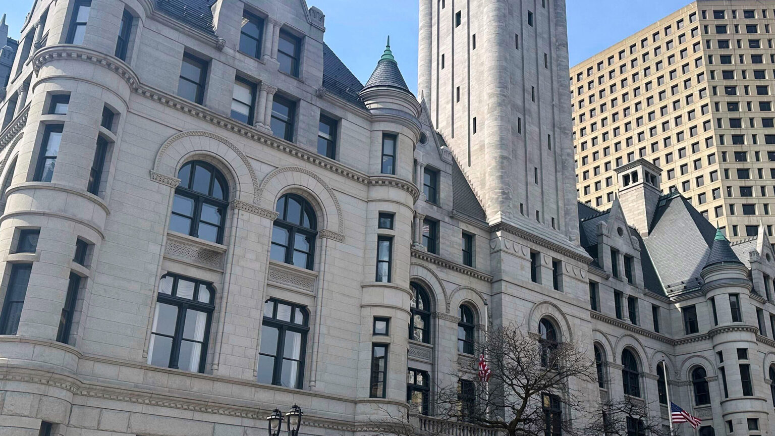 Sunlight illuminates and casts shadows on a multi-story Richardsonian Romanesque-style masonry building with multiple turrets, gables and a central tower, with two U.S. flags flying at half staff on flagpoles next to leafless trees at its central entrance and with other buildings behind it. Sunlight illuminates and casts shadows on a multi-story Richardsonian Romanesque-style masonry building with multiple turrets, gables and a central tower, with two U.S. flags flying at half staff on flagpoles next to leafless trees at its central entrance and with other buildings behind it.