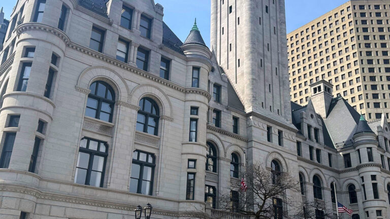 Sunlight illuminates and casts shadows on a multi-story Richardsonian Romanesque-style masonry building with multiple turrets, gables and a central tower, with two U.S. flags flying at half staff on flagpoles next to leafless trees at its central entrance and with other buildings behind it.