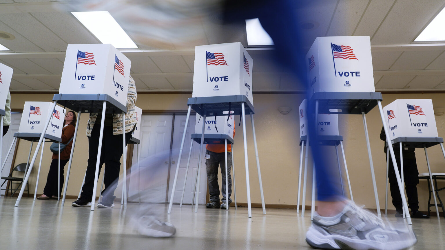 An blurry person's legs walk in front of two rows of temporary voting booths with metal legs, plastic surfaces and cardboard privacy screens with a graphic of a U.S. flag and the word Vote on each face, in a room with closed double-doors on the rear wall and fluorescent lights in a drop-panel ceiling. An blurry person's legs walk in front of two rows of temporary voting booths with metal legs, plastic surfaces and cardboard privacy screens with a graphic of a U.S. flag and the word Vote on each face, in a room with closed double-doors on the rear wall and fluorescent lights in a drop-panel ceiling.