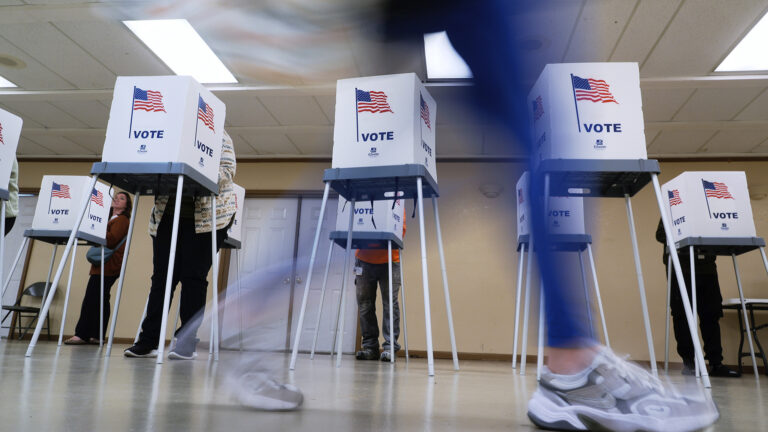 An blurry person's legs walk in front of two rows of temporary voting booths with metal legs, plastic surfaces and cardboard privacy screens with a graphic of a U.S. flag and the word Vote on each face, in a room with closed double-doors on the rear wall and fluorescent lights in a drop-panel ceiling.