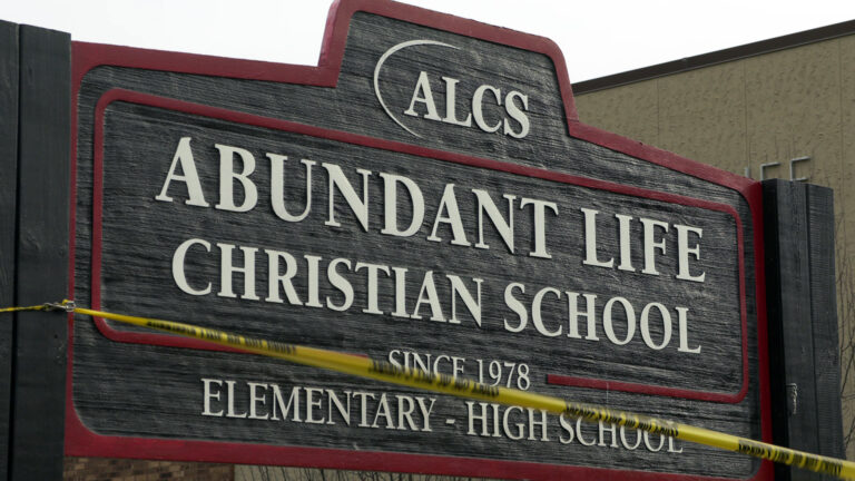 Plastic tape marked multiple times with the words Sheriff's Line Do Not Cross is attached to a wood sign with the words Abundant Life Christian School, Since 1978, Elementary - High School and a wordmark reading ALCS, with a building in the background.