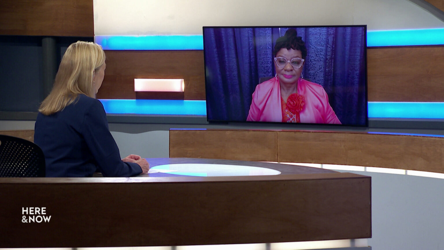Frederica Freyberg sits at a desk on the Here & Now set and faces a video monitor showing an image of Gwen Moore.
