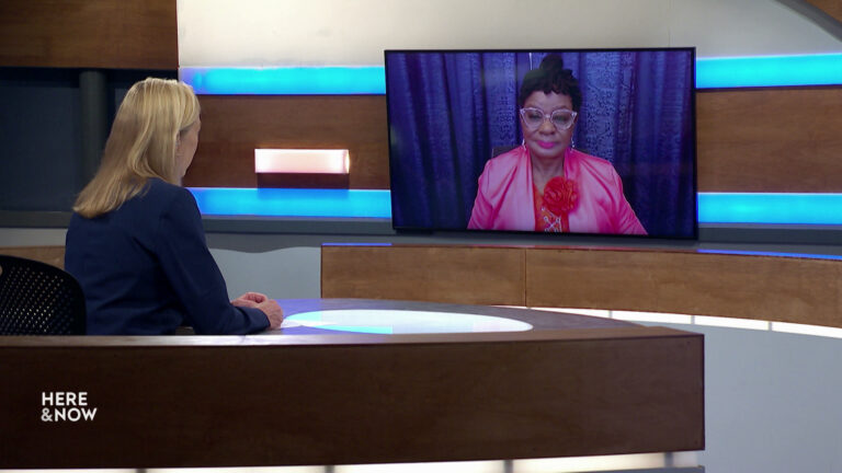 Frederica Freyberg sits at a desk on the Here & Now set and faces a video monitor showing an image of Gwen Moore.