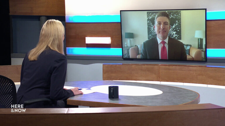 Frederica Freyberg sits at a desk on the Here & Now set and faces a video monitor showing an image of Bryan Steil.