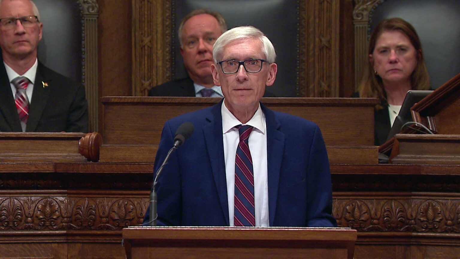 Tony Evers stands and speaks into a microphone mounted to the top of a wood podium, with three out-of-focus people seated in high-backed leather and wood chairs on a wood legislative dais in the background. Tony Evers stands and speaks into a microphone mounted to the top of a wood podium, with three out-of-focus people seated in high-backed leather and wood chairs on a wood legislative dais in the background.