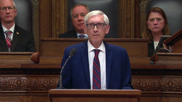 Tony Evers stands and speaks into a microphone mounted to the top of a wood podium, with three out-of-focus people seated in high-backed leather and wood chairs on a wood legislative dais in the background.