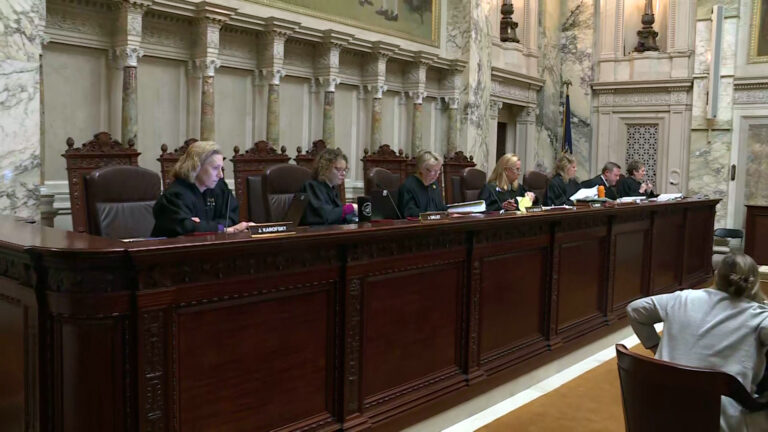 From left to right, Jill Karofsky, Rebecca Dallet, Ann Walsh Bradley, Annette Ziegler, Rebecca Bradley, Brian Hagedorn and Janet Protasiewicz sit in high-backed leather chairs at a judicial dais, with another row of high-backed wood and leather chairs behind them, in a room with marble masonry and a Wisconsin flag, with a person seated in a wood arm chair on the floor in the foreground preparing to stand.