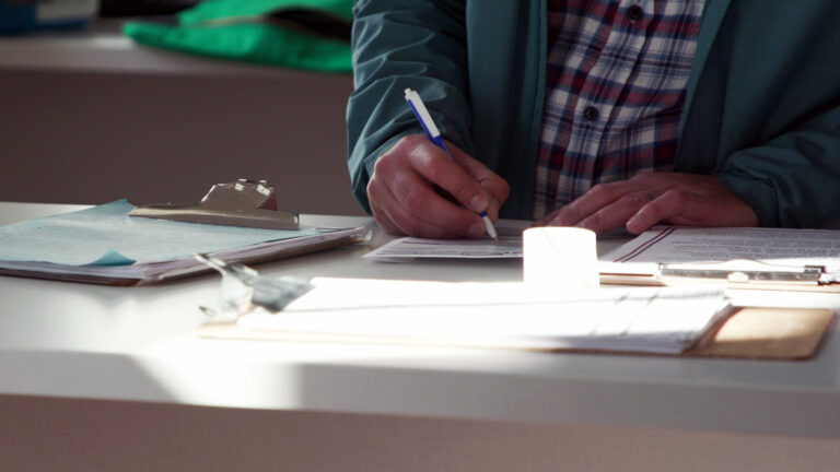 A person writes with a pen in their right hand to fill out an absentee ballot envelope, with their left hand holding its corner, while standing at a counter with multiple clipboards with papers on its surface.