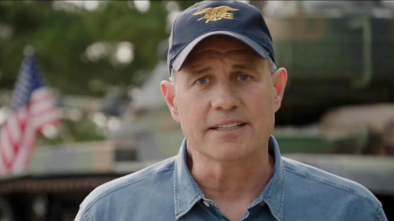 Bill Berrien speaks while standing outside with an out-of-focus U.S. flag, foliage and structures in the background.