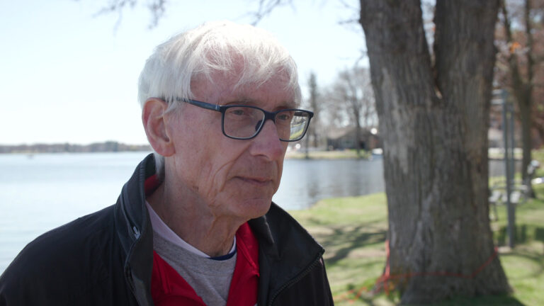 Tony Evers speaks while standing outdoor in front of a lake, with shoreline, trees, docs and buildings in the background.