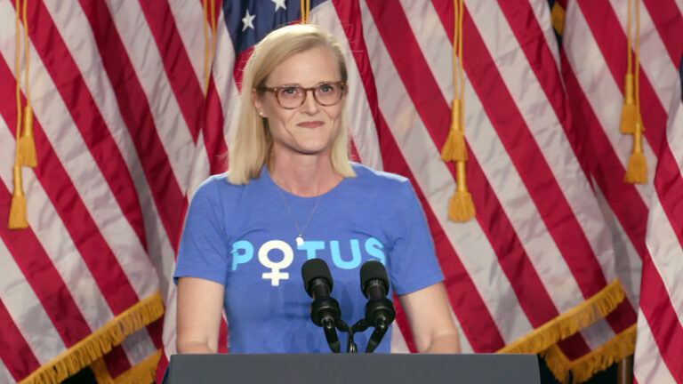 Sarah Godlewski stands and speaks into two microphones mounted to the top of a podium, with a row of U.S. flags standing behind her.