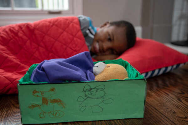 A child in a sleeping bag lays next to a stuffed animal in a handmade bed.