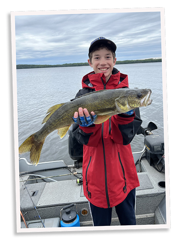 Smiling child in a red jacket and baseball cap holds a large fish on a boat, with a calm lake and forested shoreline in the background.