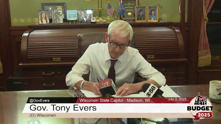 Tony Evers signs a page of a document with this right hand and with his left hand on another page, while seated at a large wood desk with a coffee mug and two microphones with the flags of different media organizations, in a room with a wood roll top desk with photos, plaques and small U.S. and Wisconsin flags on its top shelf.