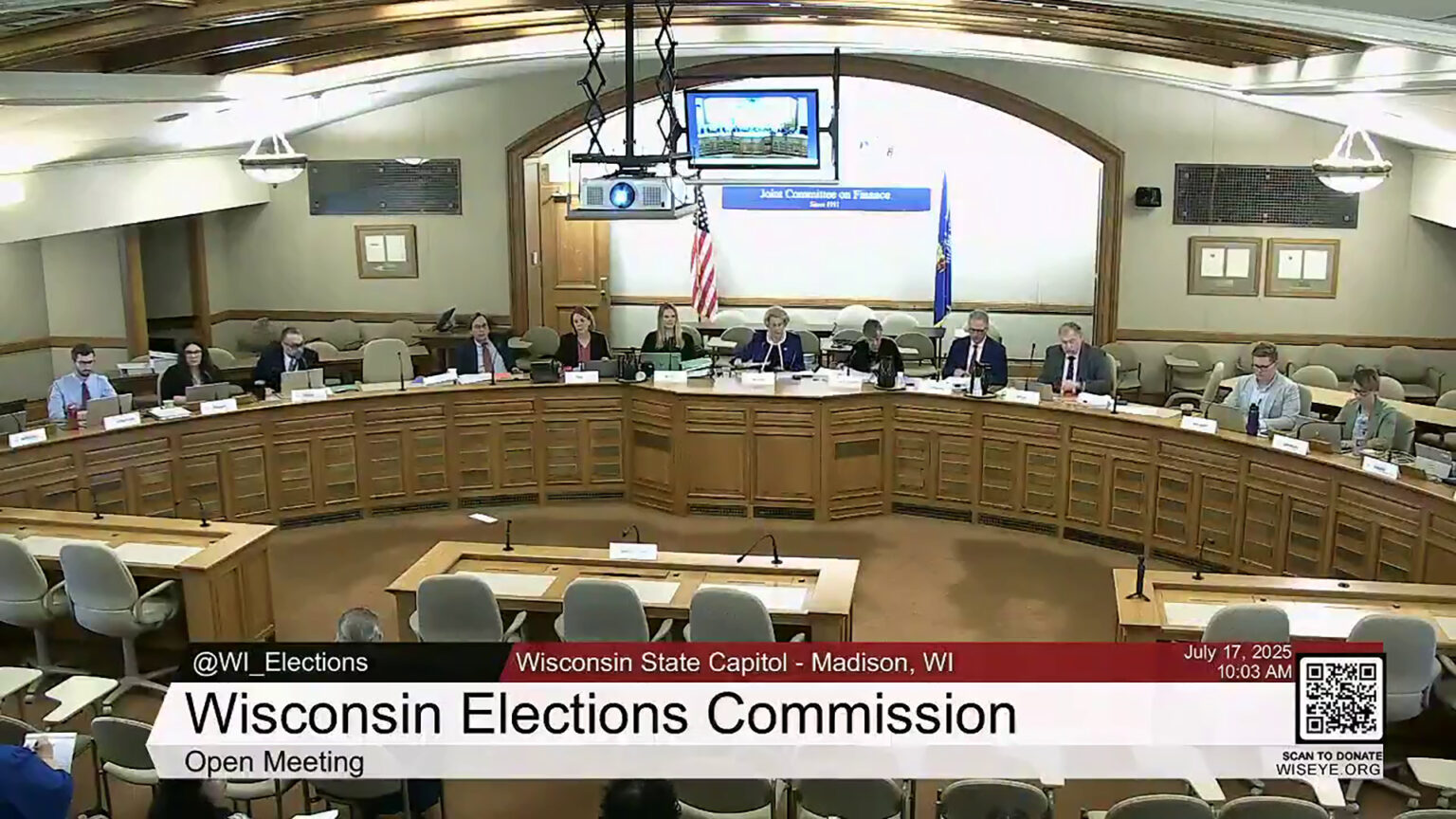 A video still image shows 12 people sitting at a legislative dais in front of microphones and nameplates, in a room with the U.S. and Wisconsin flags on either side of the Great Seal of the State of Wisconsin mounted on the rear wall, with a row of three desks and multiple rows of chairs facing the dais, with a video graphic at bottom including the text Wisconsin Elections Commission and Open Meeting.