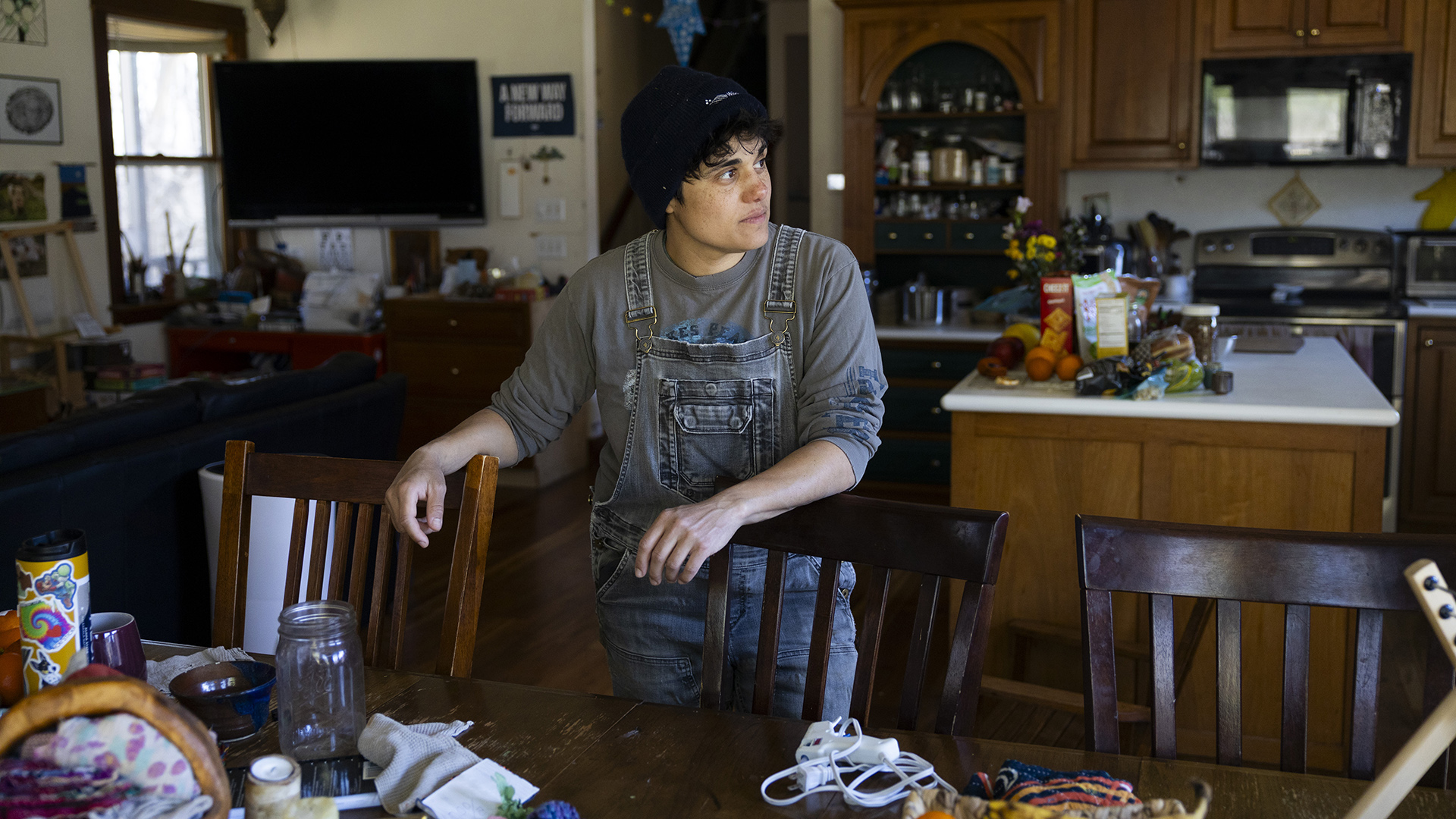Jess D'Souza stands in front of a wood dining room table with each hand over the back of a wood chair in an open-plan room, with a kitchen island, cabinets and appliances along with a desk and wall-mounted television next to a window in the background.