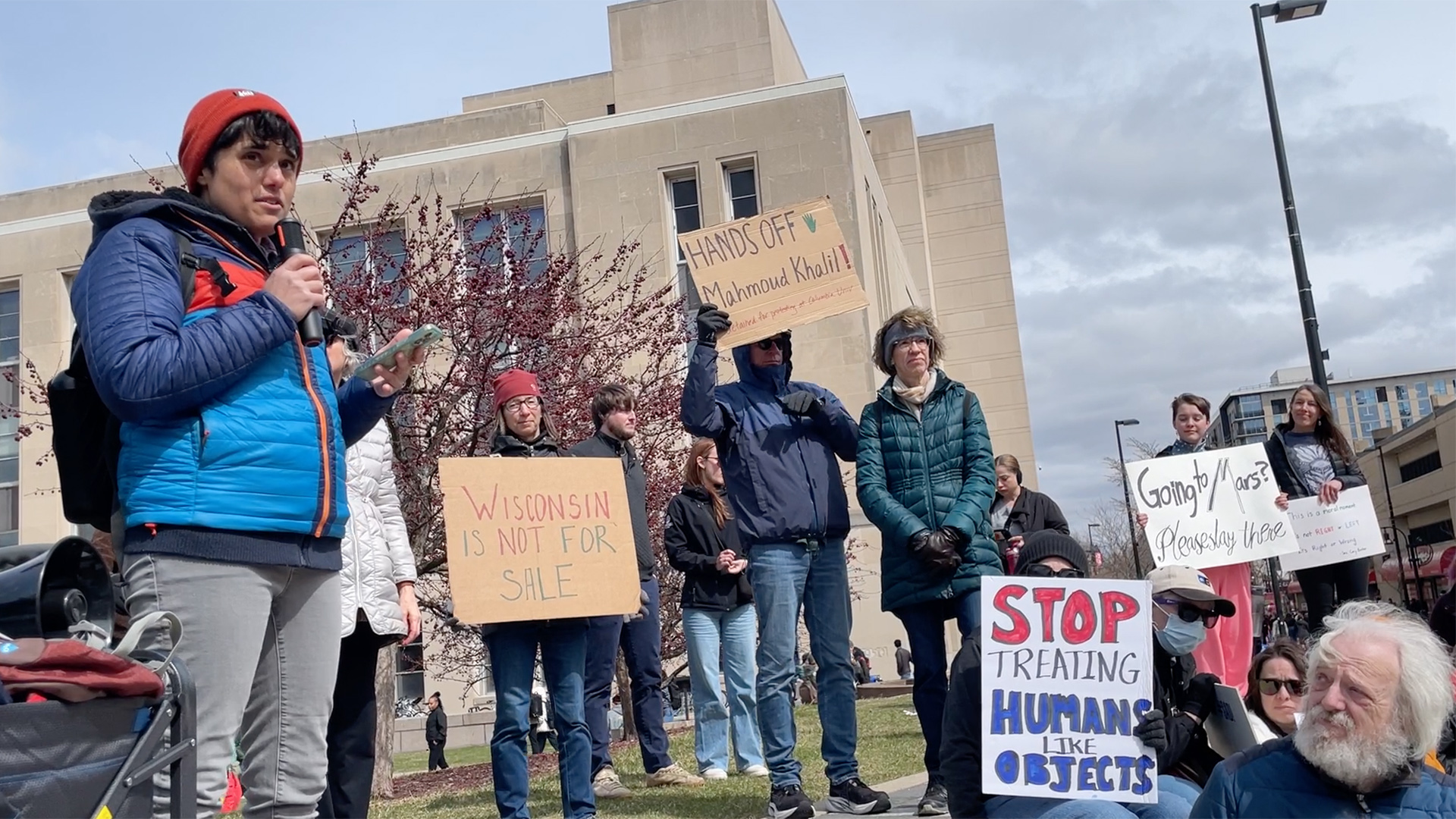 Jess D'Souza speaks into a wireless microphone in her right hand while standing next to and in front of multiple people holding hand-written signs, with multiple buildings and light poles in the background.