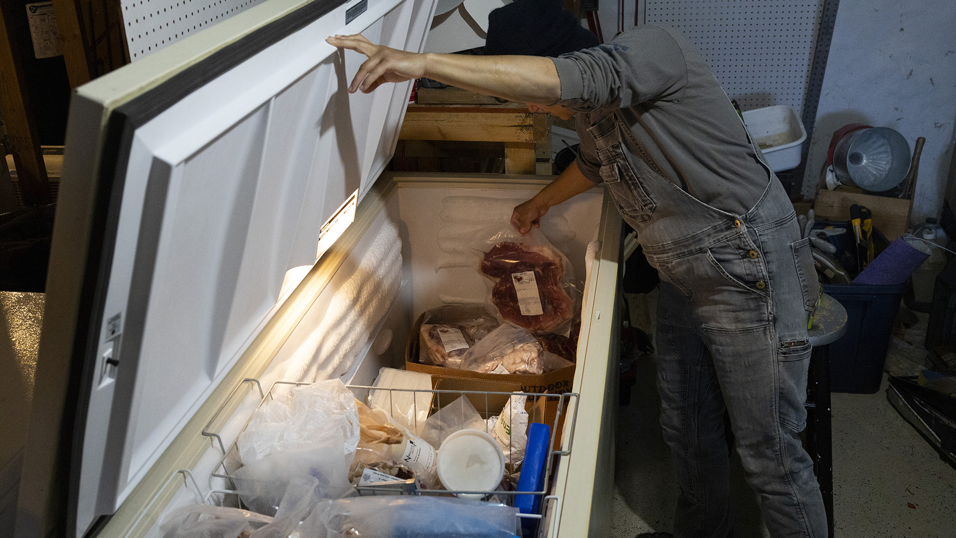 Jess D'Souza lefts a vacuum-sealed plastic package of pork from the inside of a chest freezer in a room with items stacked against a wall in the background.