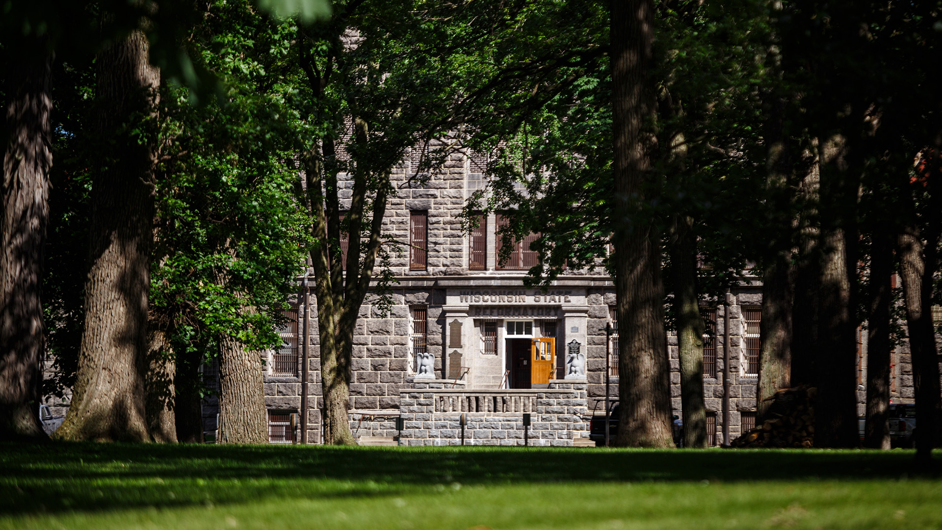 A bas-relief sign reads "Wisconsin State Reformatory" on a lintel above an open wood outer door to a multi-story stone masonry building, with a lawn and multiple large deciduous trees with full canopies in the foreground.