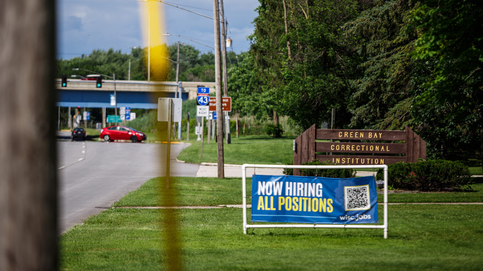 A vinyl sign with the words Now Hiring All Positions and a QR code is mounted in the middle of a PVC pipe frame that is placed on a lawn in front of a wood sign with the words Green Bay Correctional Institution, with trees, a road with vehicles, traffic signs and signals and a highway overpass in the background, and with an out-of-focus wood utility pole in the foreground. A vinyl sign with the words Now Hiring All Positions and a QR code is mounted in the middle of a PVC pipe frame that is placed on a lawn in front of a wood sign with the words Green Bay Correctional Institution, with trees, a road with vehicles, traffic signs and signals and a highway overpass in the background, and with an out-of-focus wood utility pole in the foreground.