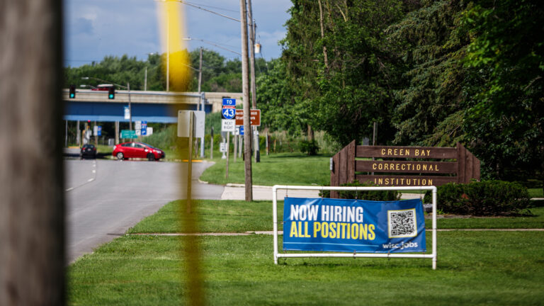 A vinyl sign with the words Now Hiring All Positions and a QR code is mounted in the middle of a PVC pipe frame that is placed on a lawn in front of a wood sign with the words Green Bay Correctional Institution, with trees, a road with vehicles, traffic signs and signals and a highway overpass in the background, and with an out-of-focus wood utility pole in the foreground.