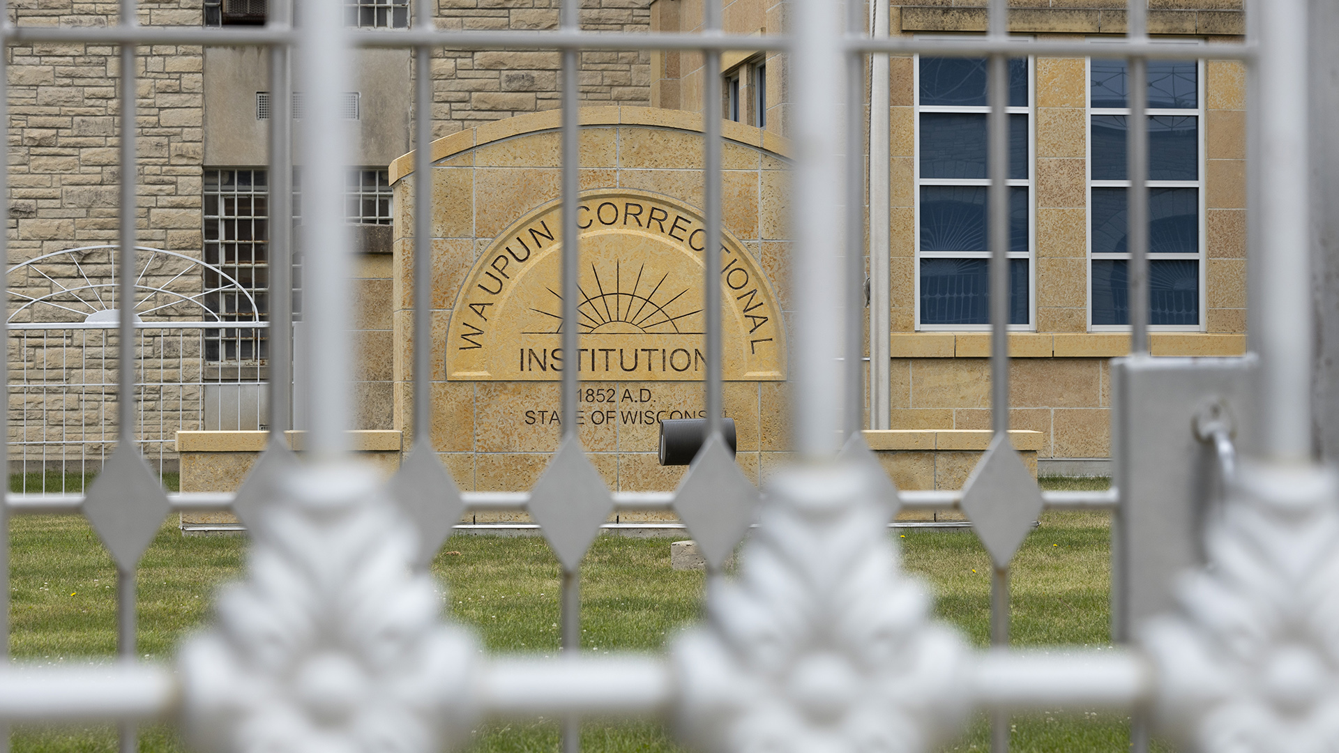 A stone masonry sign with the words "Waupun Correctional Institution," "1852 A.D." and "State of Wisconsin" along with a stylized rising/setting sun logo stands in front of a multi-story building with different types of stone masonry and windows, with an out-of-focus double set of metal fences in the foreground.