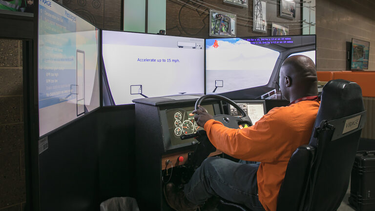 A man sits in the chair of a driving simulator and holds a steering wheel with his left hand while facing the simulator's gauge console and three computer monitors arranged in a semi-circle, in a room with framed images and wood paneling on a concrete block wall.