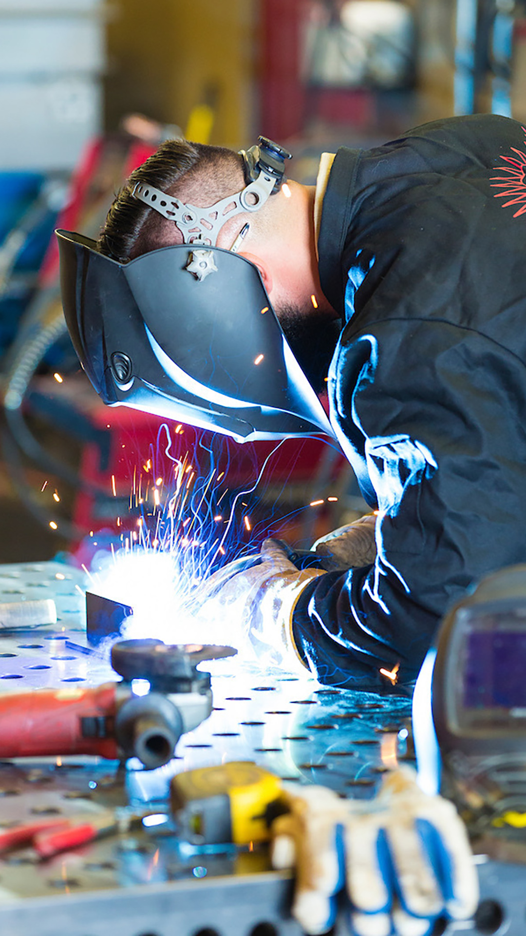 A man wearing a welding helmet leans over a welding table illuminated by a welding arc surrounded by sparks, with out-of-focus gloves, a tape measure and tools on the tables surface, in a room with out-of-focus tools in the background.