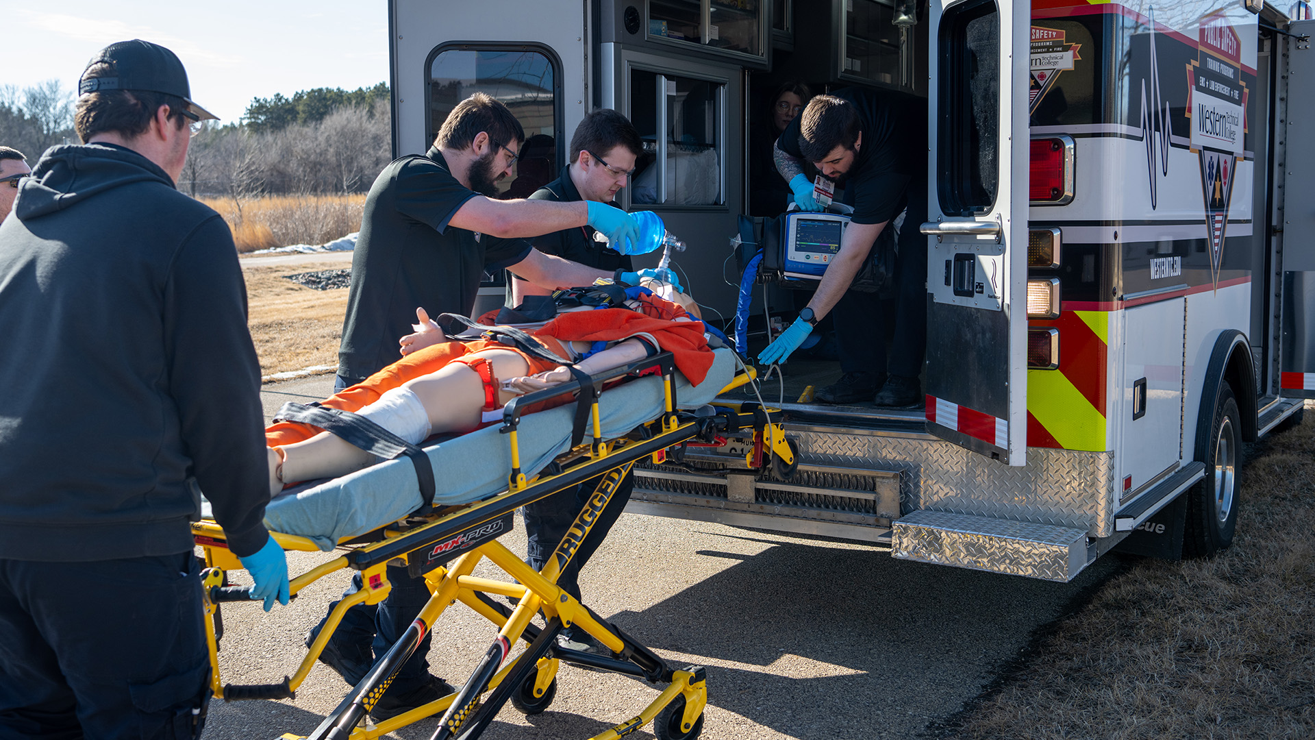 Several people stand outdoors around a folding gurney stretcher with a mannequin strapped to its surface, with one person applying a respirator to its face, next to an ambulance parked along an asphalt driveway with its rear doors open and more people seated and standing inside, with a lawn, snowbanks and leafless trees in the background.
