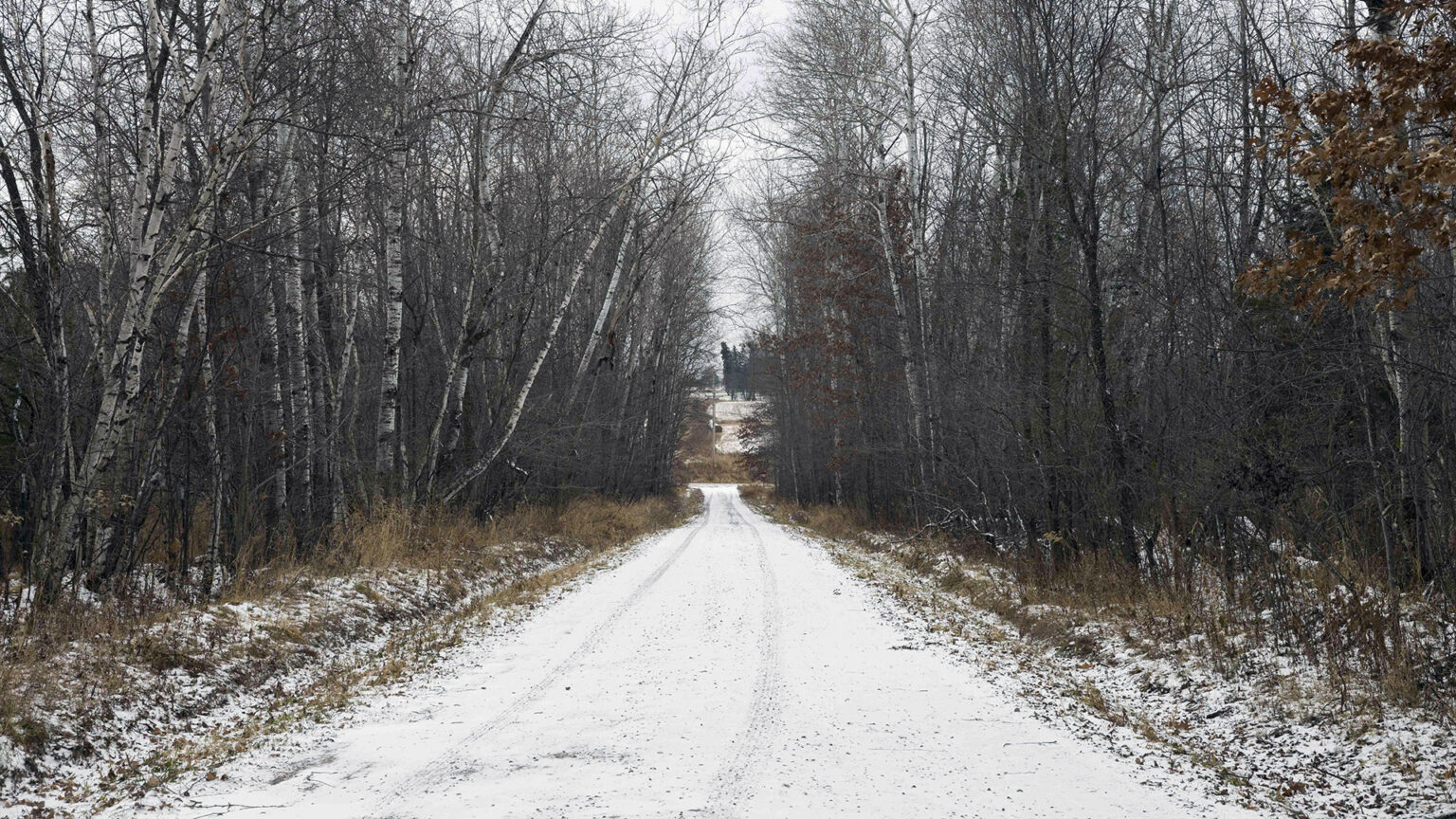 Leafless trees stand on both sides of a snow-covered gravel road with a single set of tire tracks leading toward a clearing area, with a hillside with trees on the horizon.