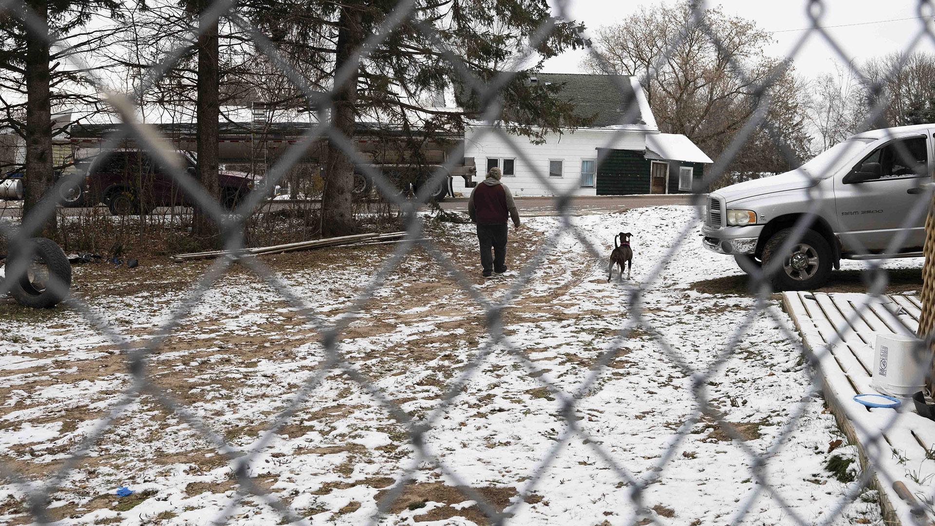Eric Zierorth and a dog walk across a gravel driveway and lawn covered by thin layer of snow and next two a snow-covered plank deck and snow-covered pickup truck, toward a SUV and tanker truck parked on a road between a stand of trees and a house, with trees in the background and an out-of-focus chain-link fence in the foreground.