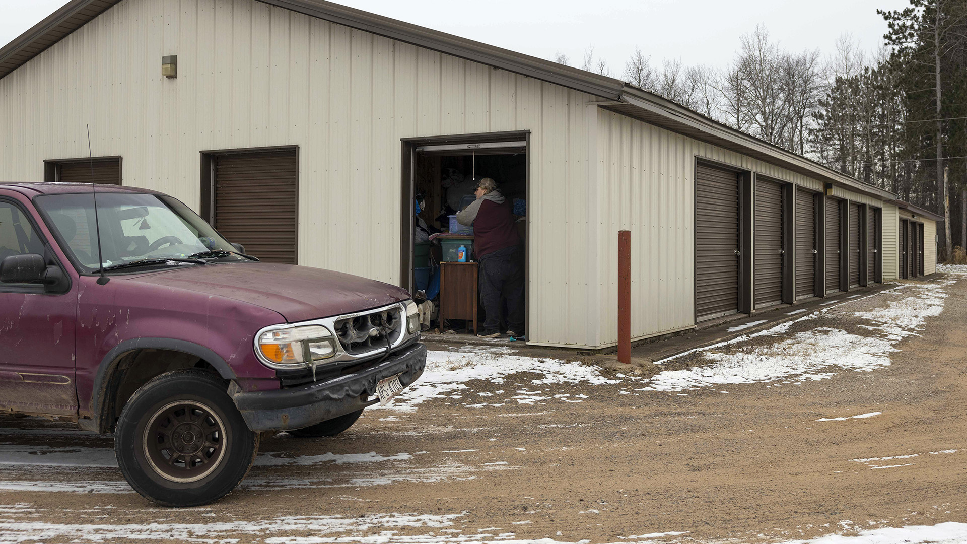 Christina Hubbell and Eric Zierorth stand on either side of a piece of furniture inside a rollup door to a storage unit in a metal building with multiple other units on each wall, with a pickup truck parked on a gravel road in the foreground and trees in the background.
