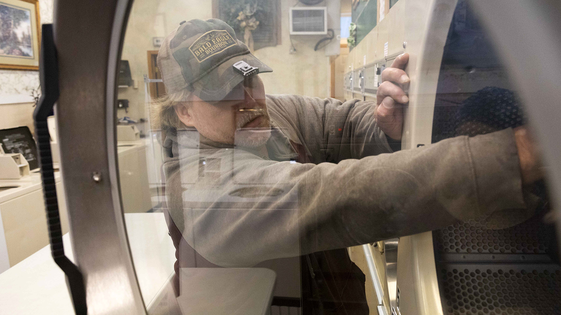 Eric Zierorth reaches inside a dryer as seen through its transparent front door while standing next to a row of driers in a laundromat, with washing machines on the opposite wall in the background.