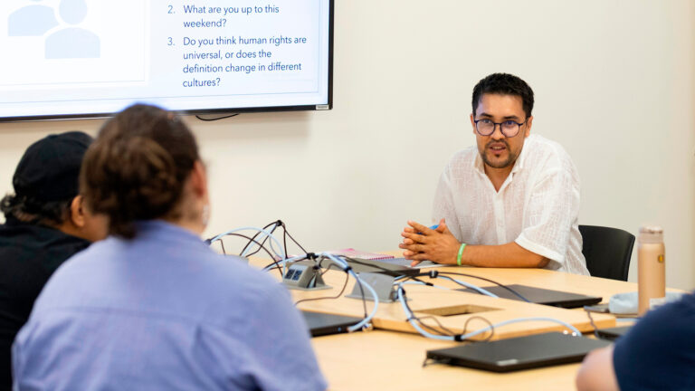 Ahmad Reca Feroz speaks with his hands folded on the surface of multiple connected tables with closed laptops and networking cords on its surface, with multiple out-of-focus people seated facing him in the foreground, with a wall-mounted monitor in the background with the words 2. What are you up to this weekend? and 3. Do you think human rights are universal, or does the definition change in different cultures? on the screen.