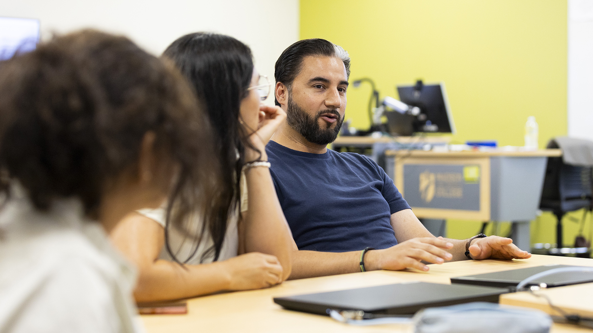 Juan Garcia speaks while seated with both hands on the surface of a table with closed laptop computers on its surface, with two people seated to his side in the foreground, and with an out-of-focus desk with a computer monitor on its surface in the background.