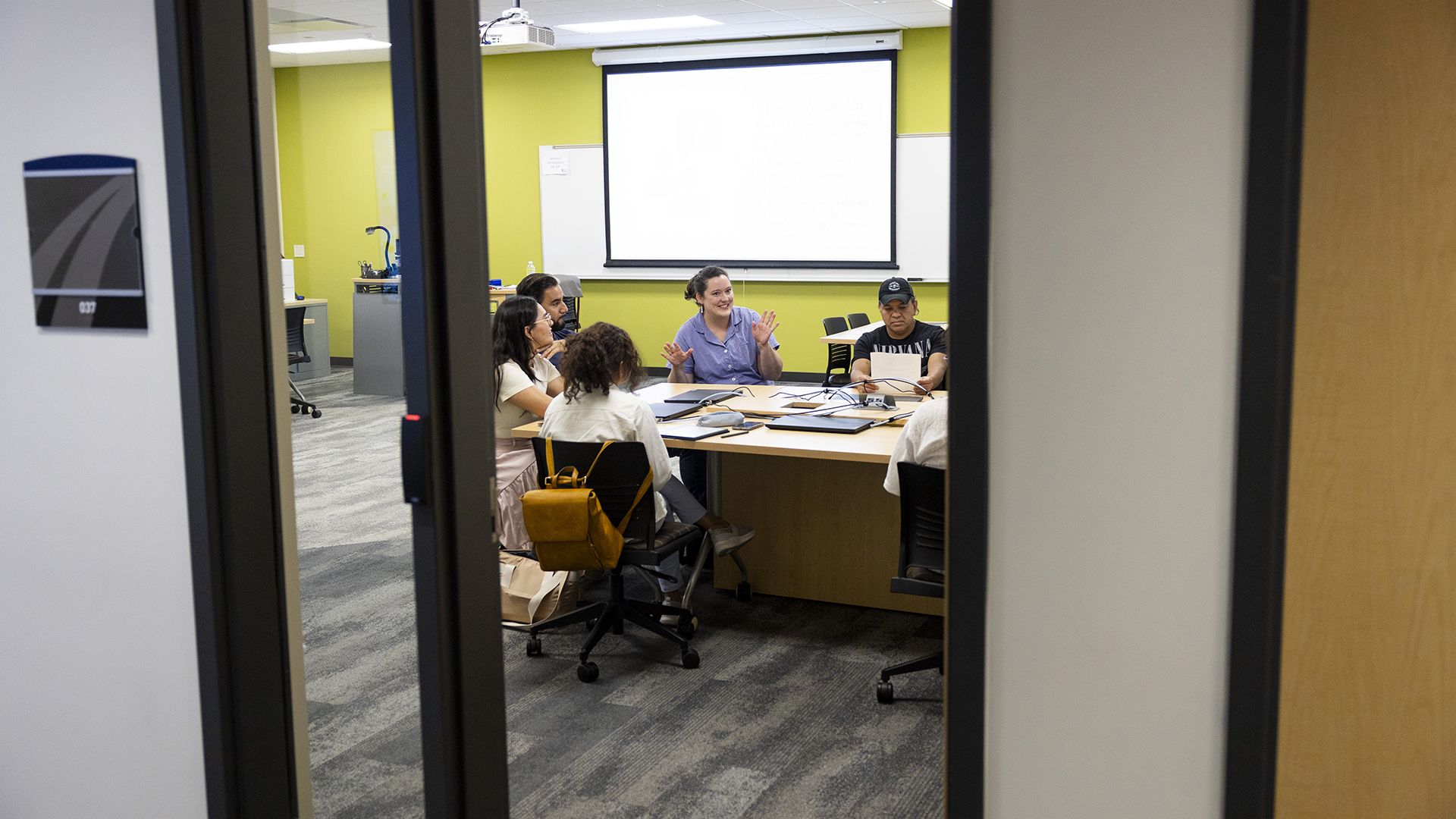 Megan Kennedy gestures with both hands while seated at a table with other people seated around her and closed laptop computers and networking cords on its surface, in a room with fluorescent overhead lighting, a low-pile carpet, and a whiteboard and lowered projector screen on the backwall, as seen through the metal and glass frame of an open doorway in the foreground.