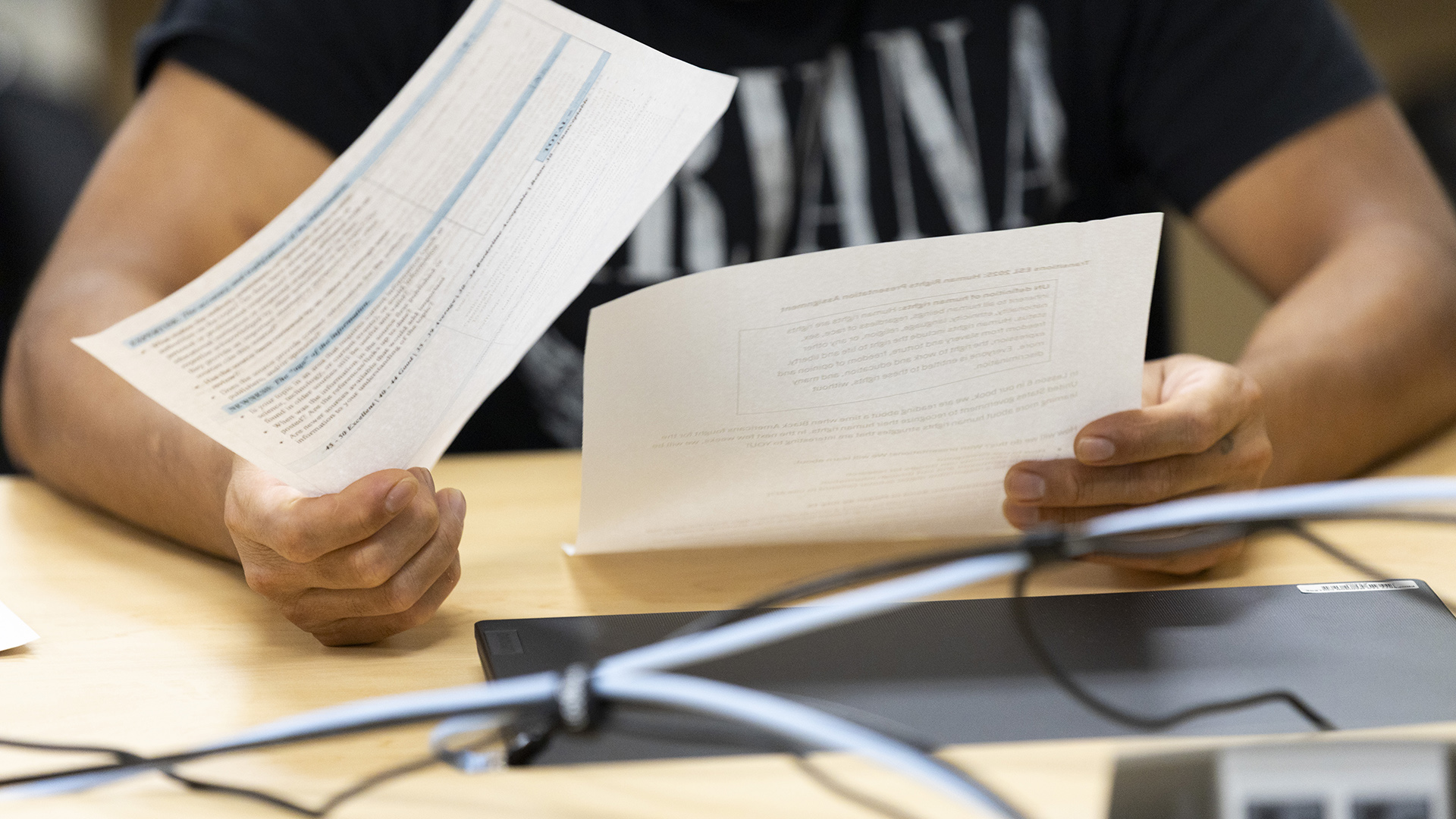 Saulo Avella Salas holds a sheet of paper with printed writing on them in each hand, while seated at a table with a closed laptop computer on its surface and out-of-focus networking cords in the foreground.