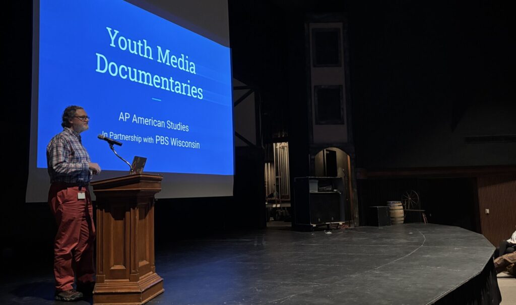 A man stands at the front of a high school auditorium, speaking to an audience.
