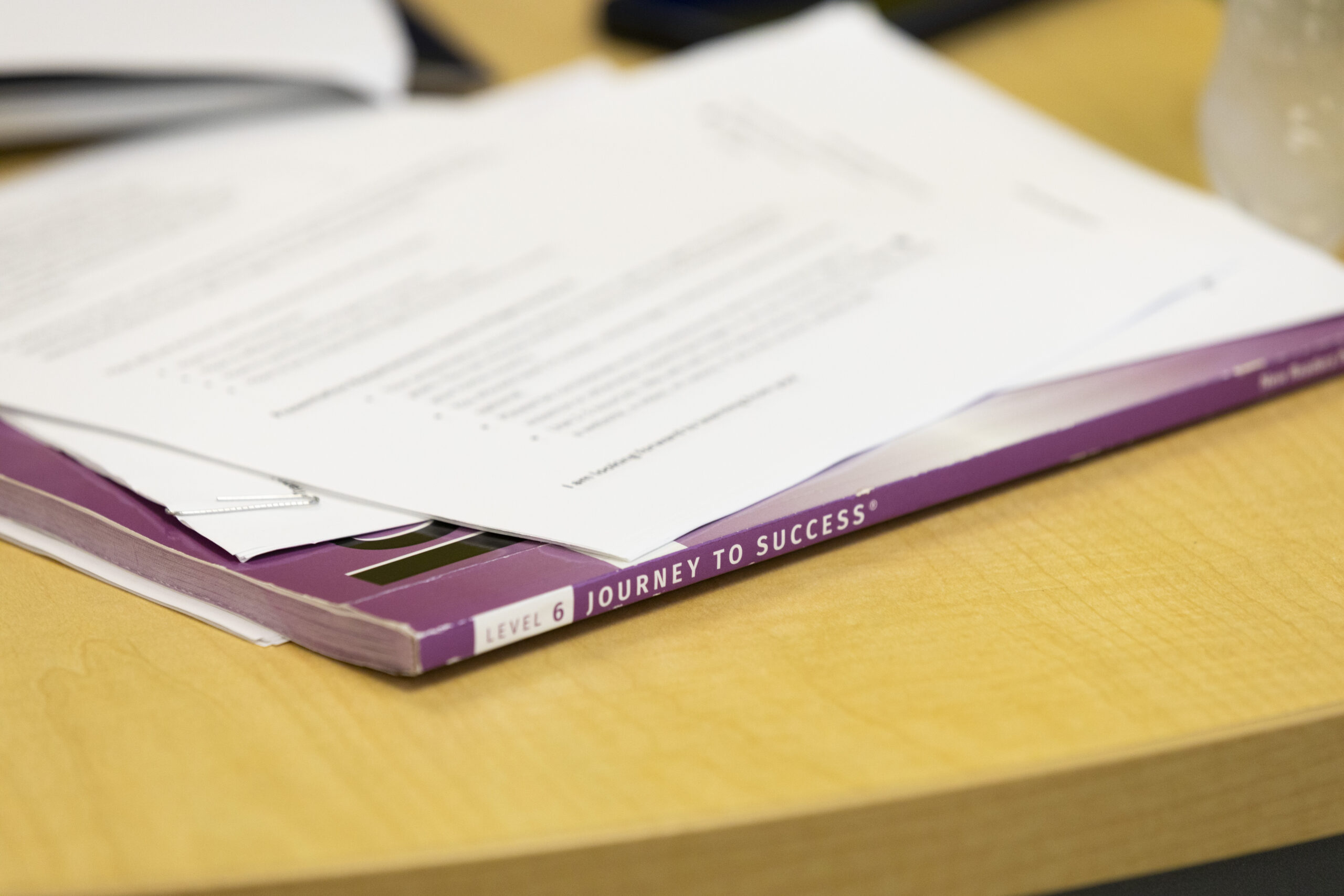 A paperbound text book with the words "Level 6" and "Journey to Success" on its spine sits on the wood-laminate surface of a table, with multiple out-of-focus papers on top of it.