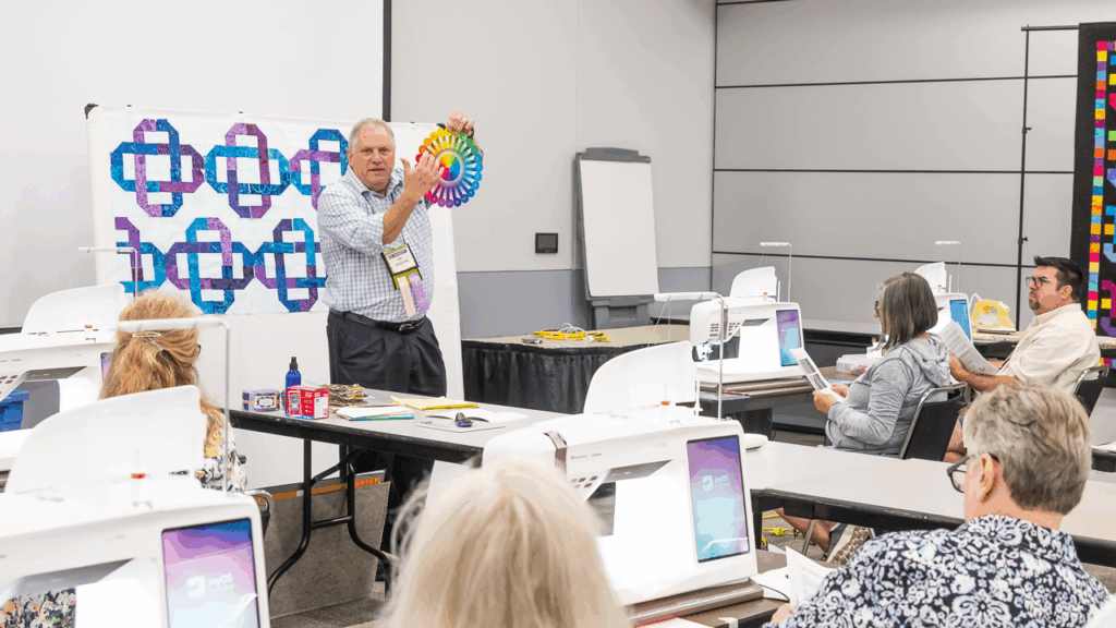 A man holds up a color wheel as he instructs a classroom of people sitting at tables, each of them have sewing machines in front of them.
