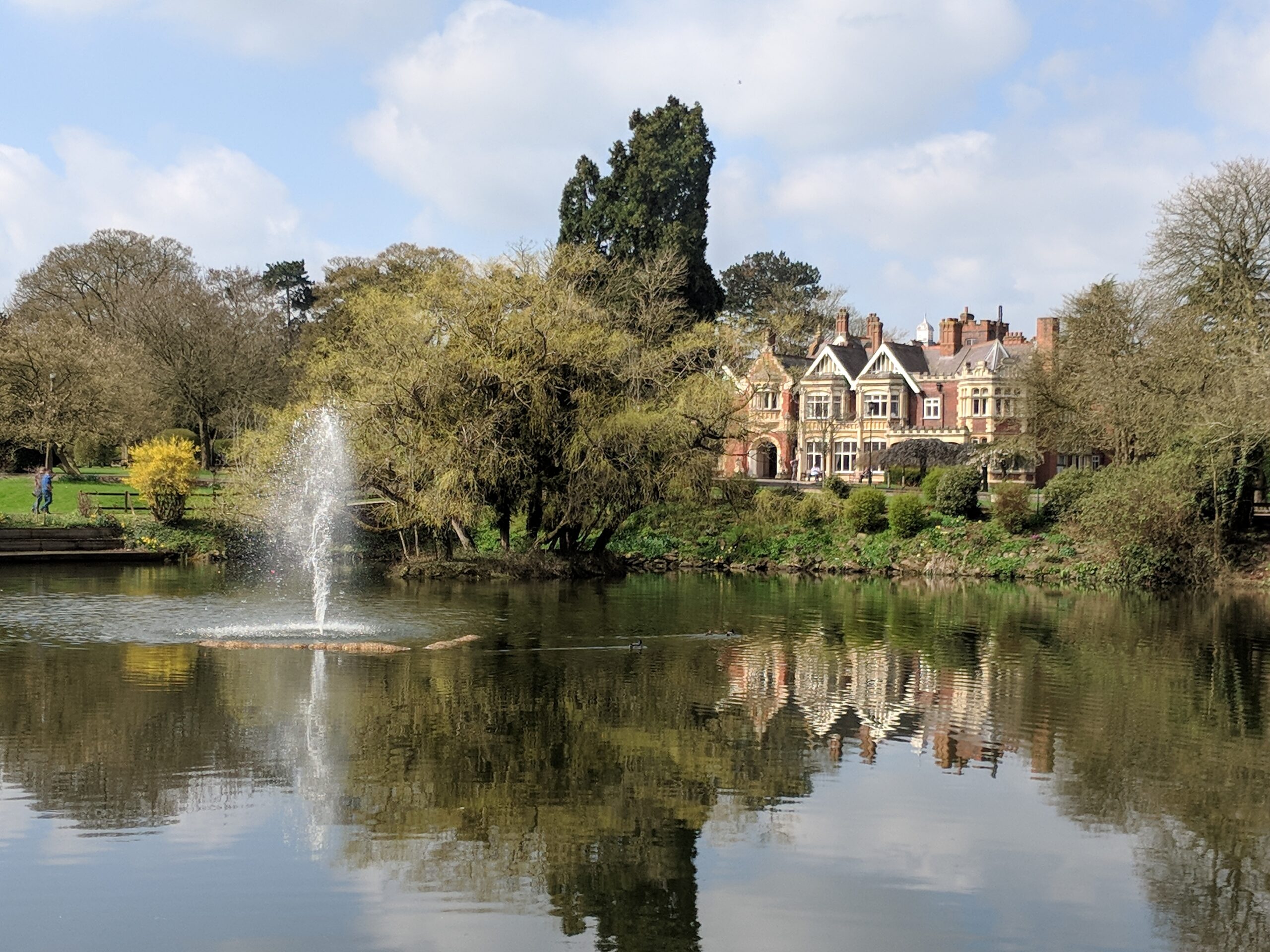 A grand historic mansion, partly hidden behind trees, with ornate architectural details and multiple chimneys. In the foreground is a calm pond with a fountain spraying water upward, surrounded by greenery and spring blooms. The sky is bright with scattered clouds, suggesting a pleasant spring day.