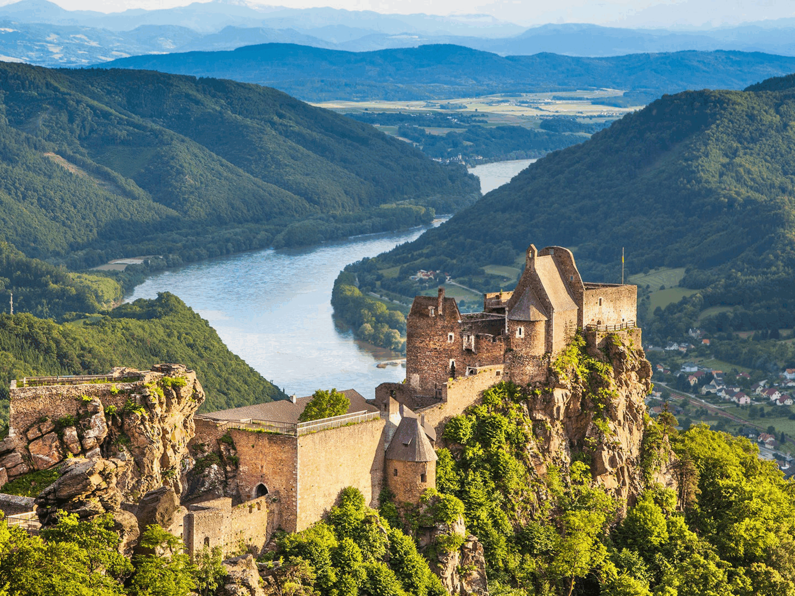 A dramatic medieval castle perched high on a rocky cliff, overlooking a winding river that snakes through a lush, green valley. The fortress, made of stone with towers and fortified walls, blends into the rugged rock formations around it. Rolling hills and forested mountains rise in the background, while a scattering of villages dot the distant landscape under a bright, clear sky.