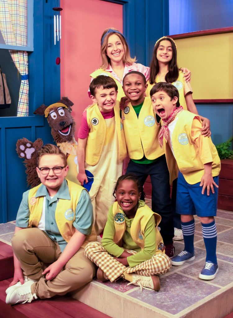 A woman, a puppet dog, and several children all smile together on a television set.
