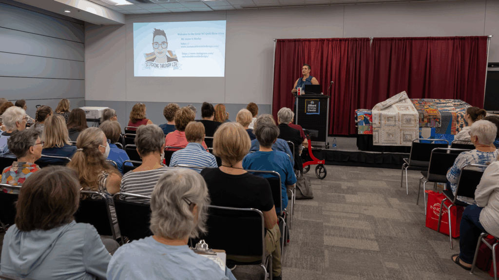 Audience listens to a speaker presenting at a quilt show, with a projection screen and colorful quilts displayed on a table beside the podium.