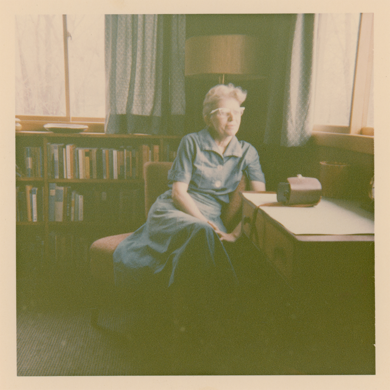 Welcome Poets - Lorine Niedecker Lorine Niedecker as an older woman in a short-sleeved dress and glasses sits in a softly lit room beside a desk and window. Bookshelves line the wall behind her, and a lamp stands between two curtained windows.