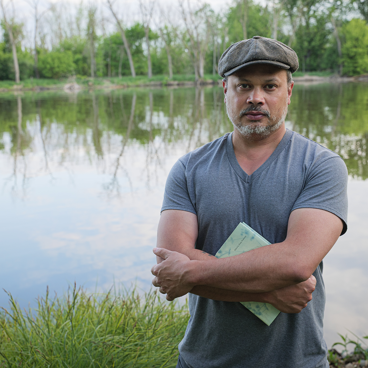 Welcome Poets - Nick Gulig Poet Nick Gulig wearing a flat cap and gray T-shirt stands on Blackhawk Island with folded arms by a calm river, holding a book against his chest. Green trees and grasses line the water behind him.