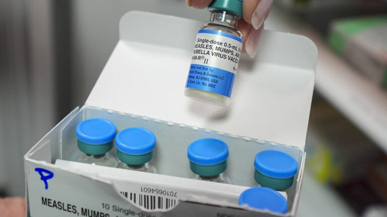 The fingertips of a left hand hold the top of a single-dose medical vile of MMR vaccine above an open paper box holding five other vials and a card with a barcode on its surface.